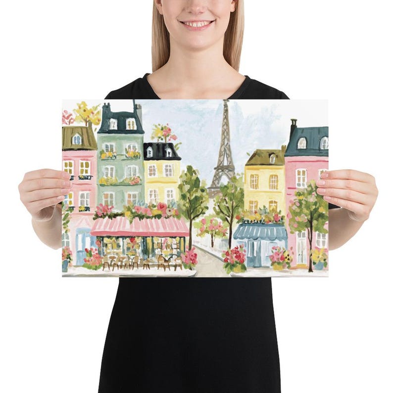 May include: A colorful print of a Parisian street scene featuring the Eiffel Tower. The artwork depicts buildings in shades of pink, green, and yellow, with flower boxes and a cafe. The print is held by a person.