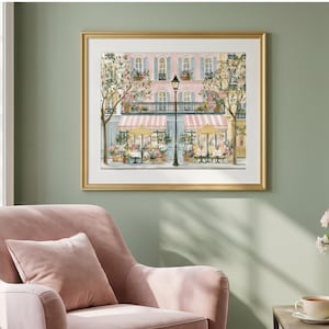 May include: A framed art print depicting a Parisian-style street scene with a pink building, striped awnings, yellow umbrellas, and a black lamppost. The illustration features potted flowers, green trees, and outdoor cafe seating in soft pastel tones.