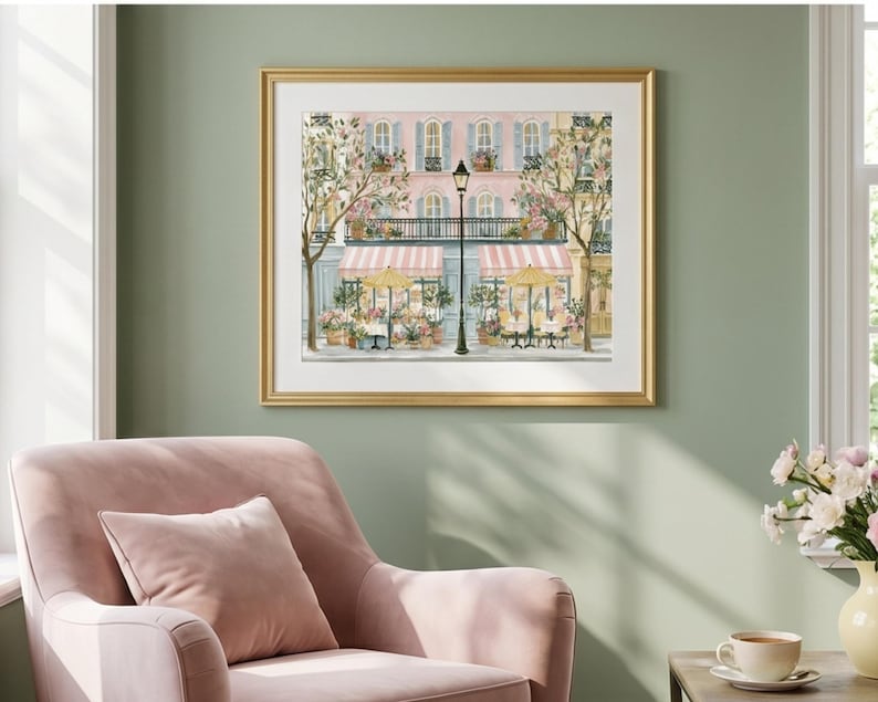 May include: A framed art print depicting a Parisian-style street scene with a pink building, striped awnings, yellow umbrellas, and a black lamppost. The illustration features potted flowers, green trees, and outdoor cafe seating in soft pastel tones.