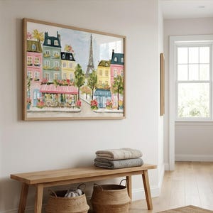 May include: A framed watercolor painting of a Parisian street scene with colorful buildings, cafes, and the Eiffel Tower. The artwork is displayed above a wooden bench with two woven baskets and folded blankets.