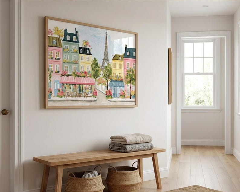 May include: A framed watercolor painting of a Parisian street scene with colorful buildings, cafes, and the Eiffel Tower. The artwork is displayed above a wooden bench with two woven baskets and folded blankets.