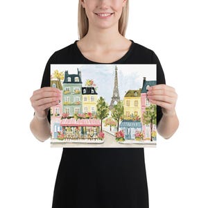 May include: Watercolor print of a Parisian street scene featuring colorful buildings, cafes, and the Eiffel Tower. The artwork showcases a charming street with floral accents and a soft, artistic style. The print is held by a person.