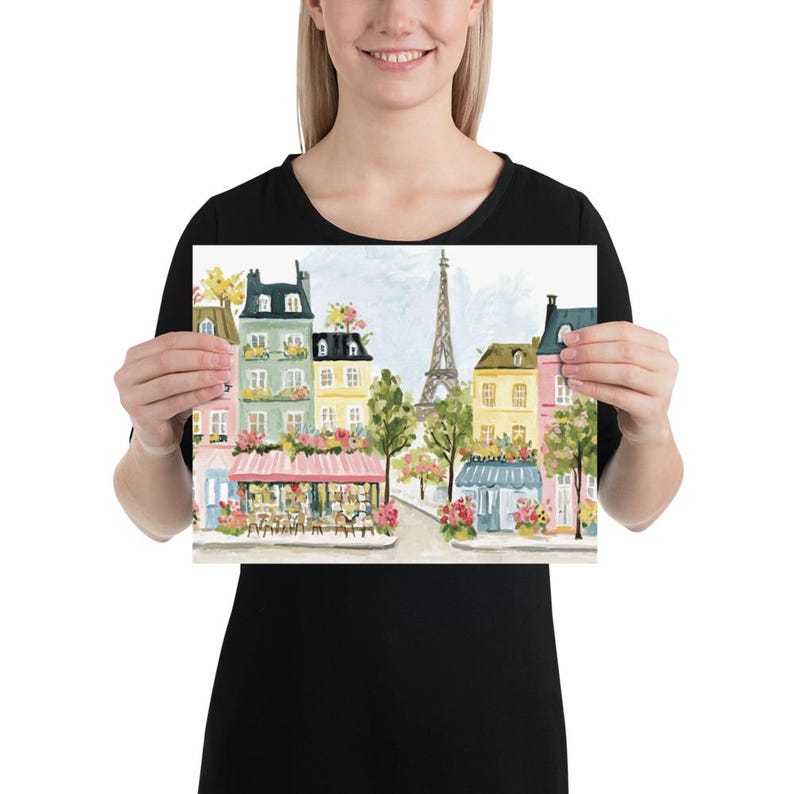 May include: Watercolor print of a Parisian street scene featuring colorful buildings, cafes, and the Eiffel Tower. The artwork showcases a charming street with floral accents and a soft, artistic style. The print is held by a person.