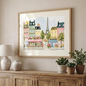 May include: A framed watercolor print of a Parisian street scene. The artwork features colorful buildings, cafes, and the Eiffel Tower. The print is in a light wood frame and is displayed above a wooden cabinet with a lamp, vase, and potted plants.
