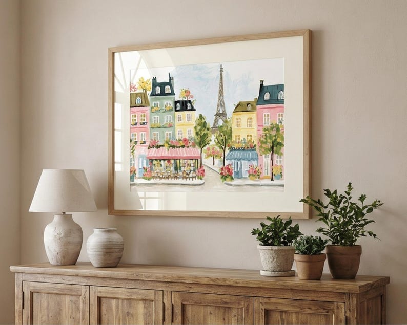 May include: A framed watercolor print of a Parisian street scene. The artwork features colorful buildings, cafes, and the Eiffel Tower. The print is in a light wood frame and is displayed above a wooden cabinet with a lamp, vase, and potted plants.