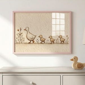 May include: A framed print featuring a mother duck and her three ducklings walking in a row. The artwork is in soft, muted tones with a light pink frame. The background is a textured beige, with a window visible.