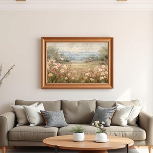 May include: A framed landscape painting with a gold-colored frame hangs above a gray sofa with decorative pillows. The painting depicts a field of pink and yellow flowers with a soft blue sky and text. A round wooden coffee table and a floor lamp complete the living room setting.
