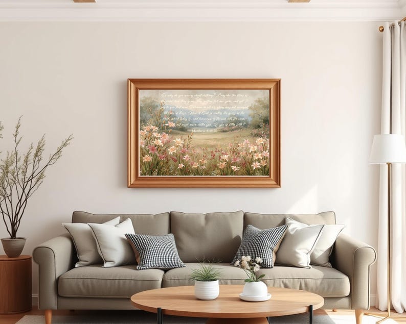 May include: A framed landscape painting with a gold-colored frame hangs above a gray sofa with decorative pillows. The painting depicts a field of pink and yellow flowers with a soft blue sky and text. A round wooden coffee table and a floor lamp complete the living room setting.