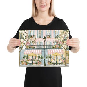 May include: A rectangular art print featuring a watercolor illustration of a Parisian-style street scene. The image displays a pink building with blue shutters, a black wrought-iron balcony, two striped pink awnings, and two yellow patio umbrellas.