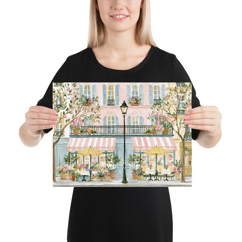 May include: A rectangular art print featuring a watercolor illustration of a Parisian-style street scene. The image displays a pink building with blue shutters, a black wrought-iron balcony, two striped pink awnings, and two yellow patio umbrellas.