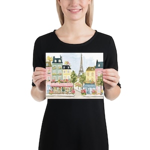 May include: A print featuring a watercolor painting of a Parisian street scene. The artwork depicts colorful buildings, cafes with pink awnings, trees, and the Eiffel Tower in the background. The overall color palette is soft and inviting.