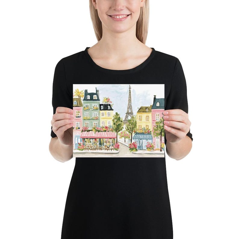 May include: A print featuring a watercolor painting of a Parisian street scene. The artwork depicts colorful buildings, cafes with pink awnings, trees, and the Eiffel Tower in the background. The overall color palette is soft and inviting.
