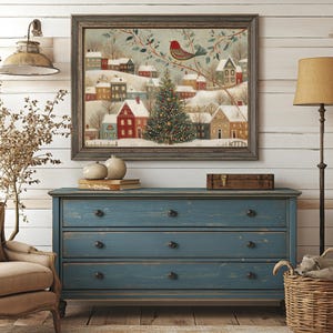 May include: A framed wall art print depicting a snowy winter village scene with colourful houses, a central decorated Christmas tree, and a red bird perched on a branch, all enclosed within a dark wooden frame with a textured finish.