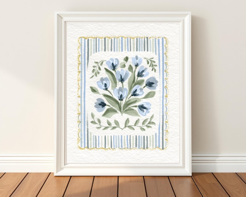 Puede incluir: Una impresi&oacute;n art&iacute;stica enmarcada que presenta un ramo de flores azules con hojas verdes. La obra de arte est&aacute; sobre un fondo blanco con un borde a rayas azules y doradas, todo dentro de un marco blanco. La impresi&oacute;n se exhibe sobre un suelo de madera.