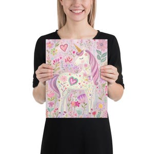 May include: A whimsical print featuring a unicorn with a golden horn and a flowing pink mane and tail. The unicorn is adorned with floral patterns and hearts, set against a backdrop of colorful flowers and foliage. The artwork is held by a person.