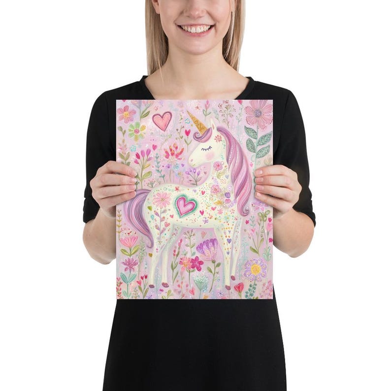 May include: A whimsical print featuring a unicorn with a golden horn and a flowing pink mane and tail. The unicorn is adorned with floral patterns and hearts, set against a backdrop of colorful flowers and foliage. The artwork is held by a person.