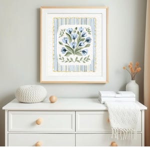 Puede incluir: Una impresi&oacute;n floral enmarcada con flores azules y hojas verdes. La obra de arte est&aacute; sobre un fondo a rayas con un borde dorado. El marco es de madera clara. La impresi&oacute;n se exhibe en una c&oacute;moda blanca.