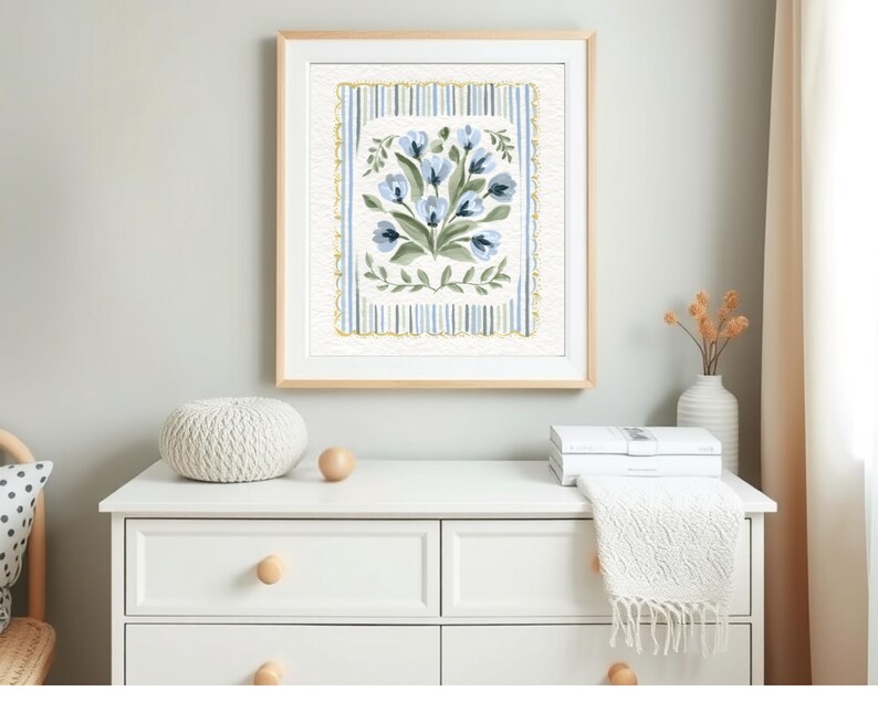 Puede incluir: Una impresi&oacute;n floral enmarcada con flores azules y hojas verdes. La obra de arte est&aacute; sobre un fondo a rayas con un borde dorado. El marco es de madera clara. La impresi&oacute;n se exhibe en una c&oacute;moda blanca.