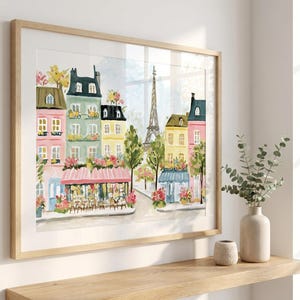 May include: Watercolor painting of a Parisian street scene with colorful buildings, cafes, and the Eiffel Tower. The artwork is framed in a light wood frame and displayed on a wooden shelf. A vase with greenery and a small decorative pot are on the shelf.