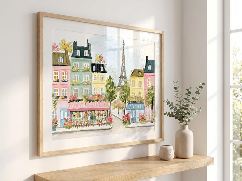 May include: Watercolor painting of a Parisian street scene with colorful buildings, cafes, and the Eiffel Tower. The artwork is framed in a light wood frame and displayed on a wooden shelf. A vase with greenery and a small decorative pot are on the shelf.