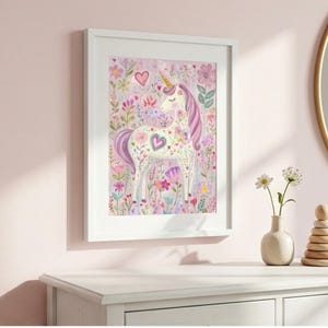 May include: A framed watercolor print of a unicorn surrounded by flowers in shades of pink, purple, and green. The unicorn has a golden horn and a heart-shaped design on its body. The artwork is displayed on a white dresser.