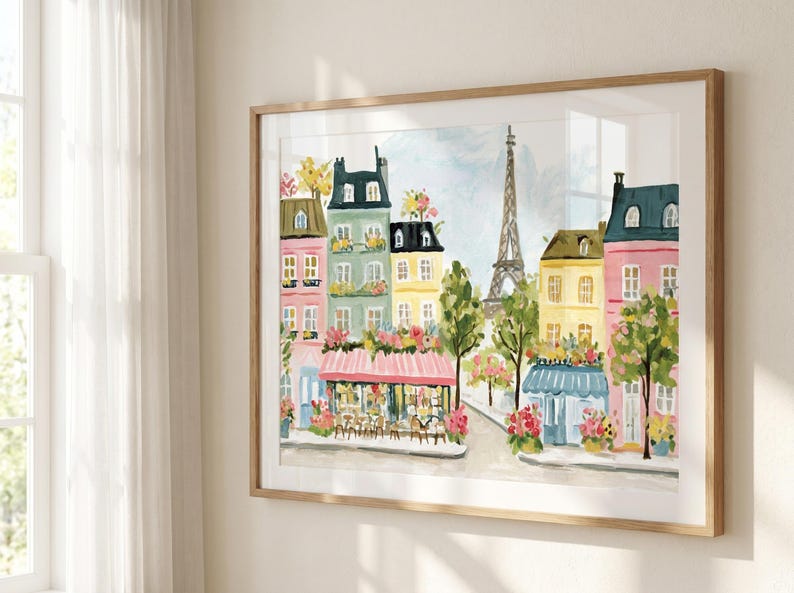May include: Watercolor painting of a Parisian street scene with colorful buildings, cafes, and the Eiffel Tower. The artwork is framed in a light wood frame and hangs on a white wall. The buildings are painted in pastel colors.