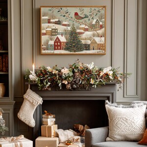 May include: A rectangular framed wall art print featuring a winter village scene with snow-covered houses, a central Christmas tree, and a red bird perched on a branch. The artwork is displayed above a fireplace mantel decorated with greenery.