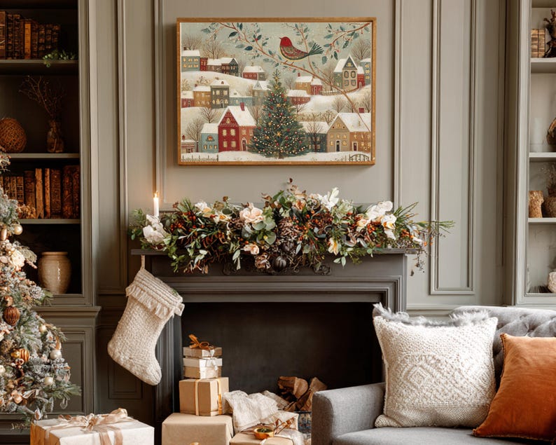 May include: A rectangular framed wall art print featuring a winter village scene with snow-covered houses, a central Christmas tree, and a red bird perched on a branch. The artwork is displayed above a fireplace mantel decorated with greenery.