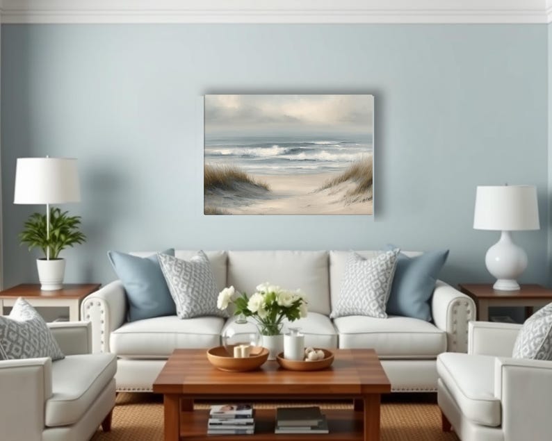 May include: A living room scene with a light blue wall, white sofa, and a wooden coffee table. A beach landscape painting hangs above the sofa. Two white lamps sit on side tables. Decorative pillows and a vase of flowers add to the decor.