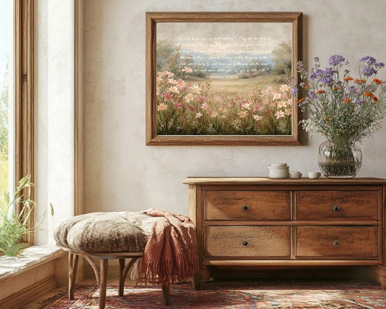 May include: A framed landscape print with a field of pink and white flowers, and text from Matthew 6:28-30. The print hangs above a wooden dresser. A bench with a patterned cushion and a terracotta throw blanket is in front of the dresser.