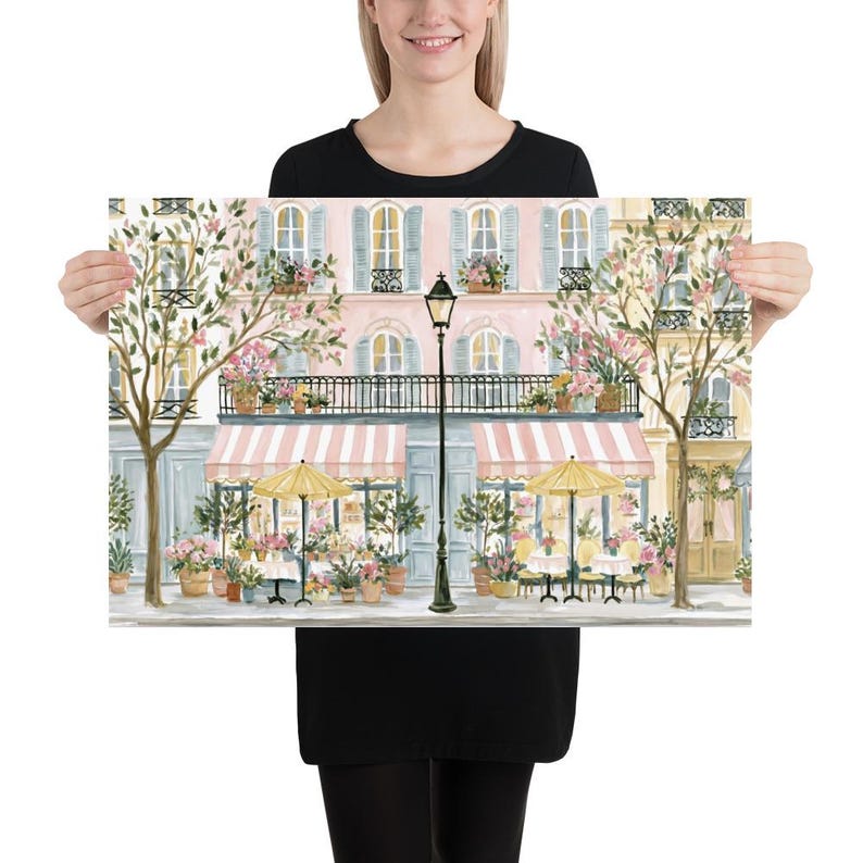 May include: A rectangular art print depicting a Parisian-style street scene with a pink building, a black lamppost, striped awnings, and yellow umbrellas over outdoor cafe tables. The scene includes potted flowers, trees, and windows with shutters.