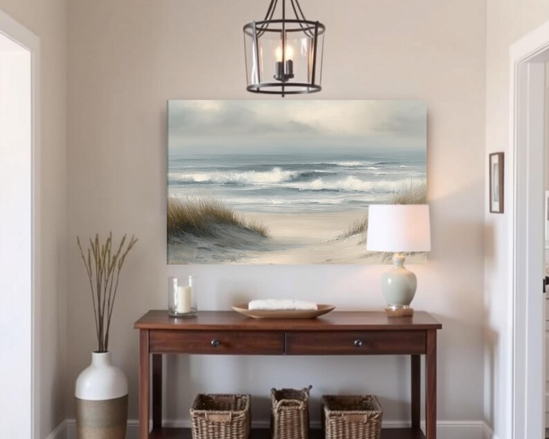 May include: A coastal-themed interior scene featuring a framed seascape painting above a dark wood console table. The painting depicts ocean waves and a sandy beach. A white lamp, decorative vase, and woven baskets complete the decor.