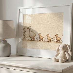 May include: A framed print of a mother duck and three ducklings walking in a row, with a neutral color palette. The artwork is displayed on a white dresser, next to a gray lamp and a stuffed bunny.