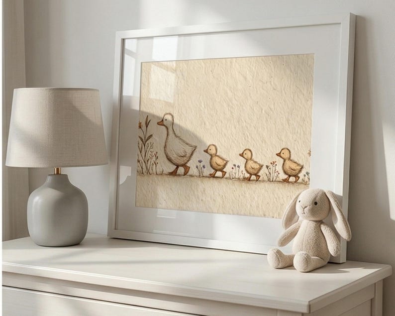 May include: A framed print of a mother duck and three ducklings walking in a row, with a neutral color palette. The artwork is displayed on a white dresser, next to a gray lamp and a stuffed bunny.