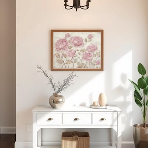 May include: A white console table with three drawers and brass knobs. A framed floral print with pink roses hangs above the table. A silver vase with branches, a decorative egg, and a potted plant complete the decor.