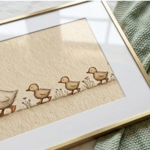 May include: A framed art print featuring a mother duck and three ducklings walking in a line. The illustration is in soft brown tones with delicate floral accents. The frame is gold-colored, and the background is a light beige.