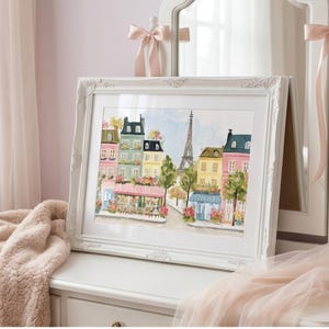 May include: A framed watercolor painting of a Parisian street scene. The artwork features colorful buildings, a cafe, and the Eiffel Tower. The white frame has ornate detailing and is displayed on a white dresser. Soft pink accents and a fluffy blanket add to the aesthetic.