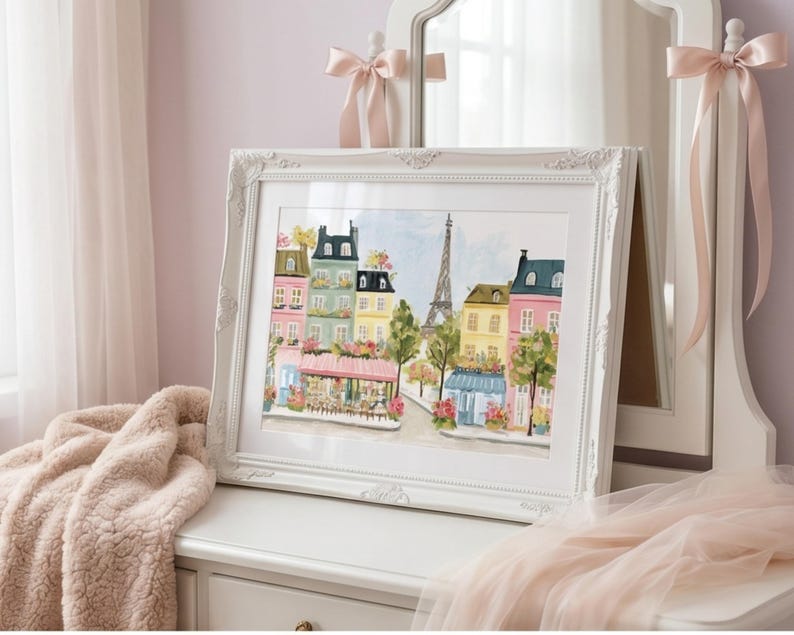 May include: A framed watercolor painting of a Parisian street scene. The artwork features colorful buildings, a cafe, and the Eiffel Tower. The white frame has ornate detailing and is displayed on a white dresser. Soft pink accents and a fluffy blanket add to the aesthetic.