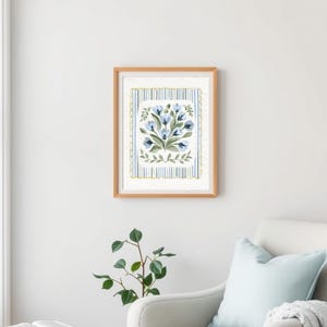 Puede incluir: Una impresi&oacute;n en acuarela enmarcada que presenta un ramo de flores azules con hojas verdes. La obra de arte est&aacute; sobre un fondo a rayas con detalles en azul y dorado, dentro de un marco de madera marr&oacute;n claro. La impresi&oacute;n se exhibe en una pared blanca.