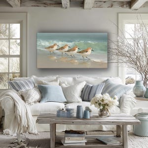 May include: A coastal-themed living room features a white sofa with blue and white pillows and a painting of shorebirds on the wall. A rustic wooden coffee table holds candles and books, with a vase of white flowers.