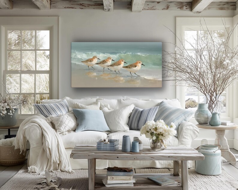 May include: A coastal-themed living room features a white sofa with blue and white pillows and a painting of shorebirds on the wall. A rustic wooden coffee table holds candles and books, with a vase of white flowers.