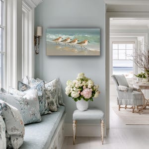 May include: An oil painting depicts three sandpipers walking along a sandy beach with gentle waves. The artwork is displayed on a light blue wall in a bright, airy room with a window seat and floral pillows.