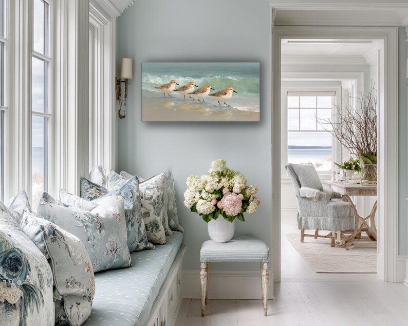 May include: An oil painting depicts three sandpipers walking along a sandy beach with gentle waves. The artwork is displayed on a light blue wall in a bright, airy room with a window seat and floral pillows.