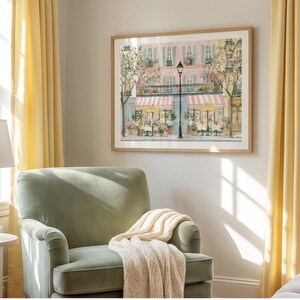 May include: A framed wall art print depicting a Parisian-style street scene with a pink building, striped awnings, outdoor cafe tables, and a tall black lamppost. The artwork features soft pastel tones, flowering trees, and architectural details.