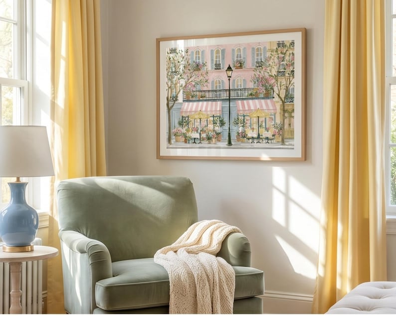May include: A framed wall art print depicting a Parisian-style street scene with a pink building, striped awnings, outdoor cafe tables, and a tall black lamppost. The artwork features soft pastel tones, flowering trees, and architectural details.