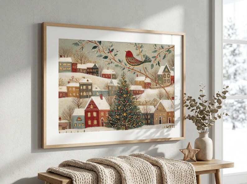 May include: A framed horizontal print depicting a snowy winter village scene with colourful houses, a large decorated Christmas tree in the centre, and a red bird perched on a branch above. The artwork features soft muted tones and festive details.