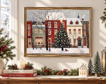 Puede incluir: Una impresión de arte mural enmarcada que representa una plaza de pueblo nevada con edificios rojos y crema, un gran árbol de Navidad decorado con una estrella, una farola y un pájaro cardenal rojo en el suelo bajo la nieve.