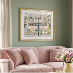 May include: A framed wall art print depicting a Parisian street scene with a pink building, striped awnings, outdoor cafe tables, yellow umbrellas, and a black street lamp, all rendered in a soft color palette with floral accents.