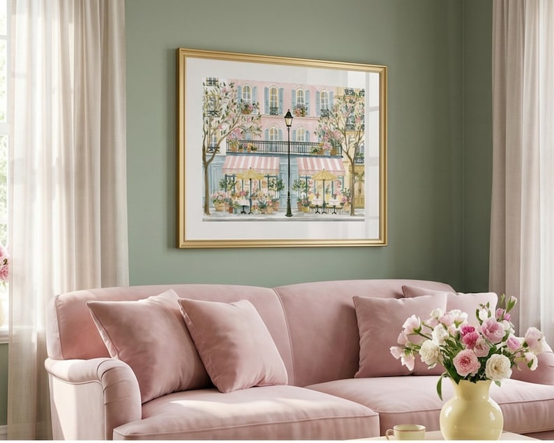 May include: A framed wall art print depicting a Parisian street scene with a pink building, striped awnings, outdoor cafe tables, yellow umbrellas, and a black street lamp, all rendered in a soft color palette with floral accents.