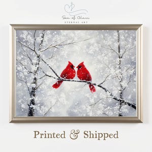 May include: A framed painting depicts two bright red cardinals perched on a snow-covered tree branch. The artwork features a winter landscape with white snow and bare trees. The painting is in a silver frame, with the text "Sea of Glass Eternal Art" at the top.